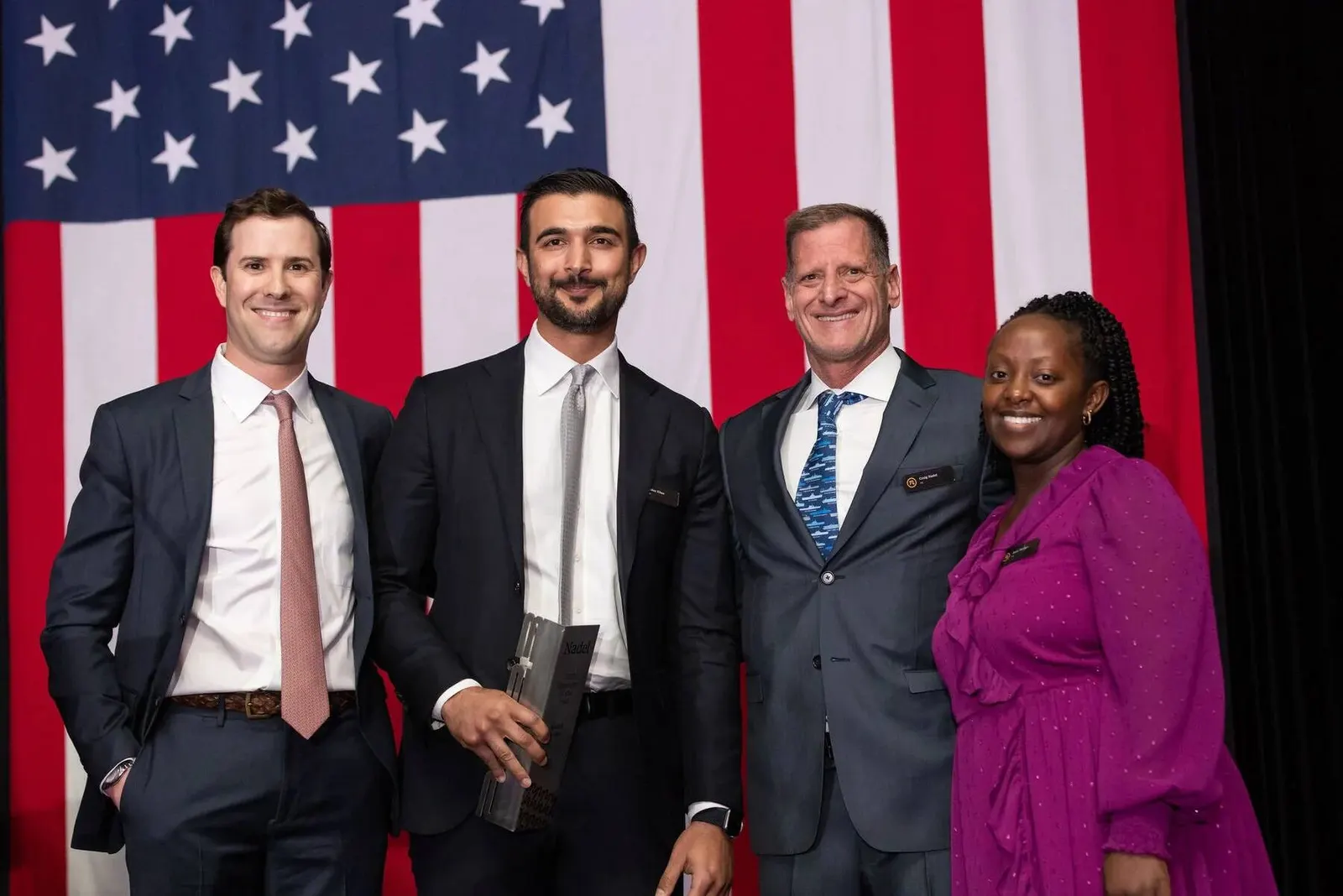 Four professionals posing together in front of an American flag backdrop at the Baber Khan Award event, photographed by Marquee Visuals.