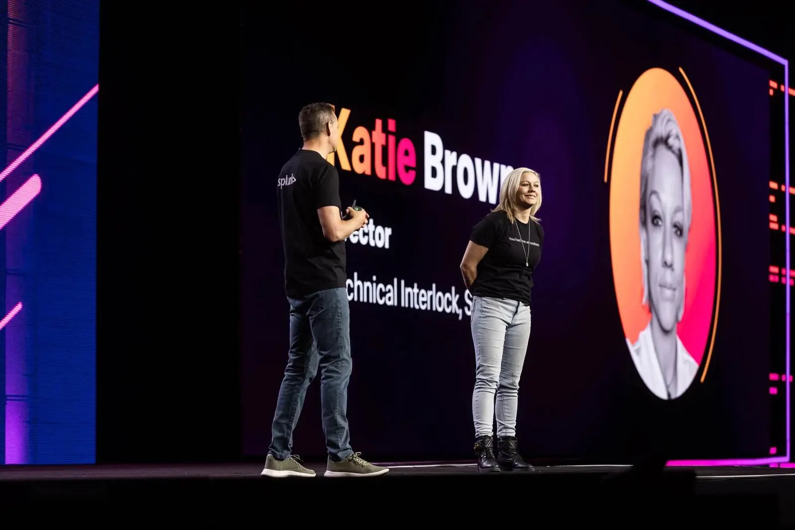 Two presenters on a conference stage with a speaker headshot and bio graphic displayed on the screen behind them, by Marquee Visuals.