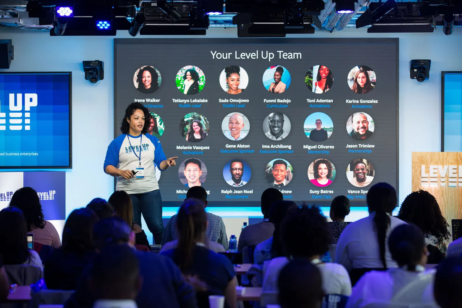 Speaker presenting on stage to a seated audience in front of a Level Up branded screen with team photos, by Marquee Visuals.