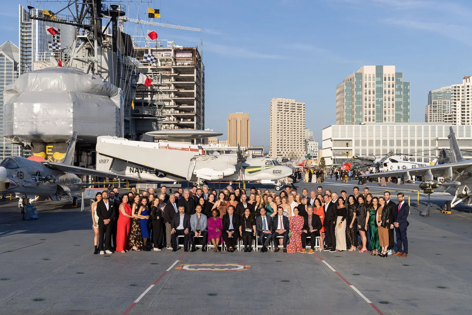 Large group photo of conference attendees on an aircraft carrier deck with a city skyline in the background, by Marquee Visuals.