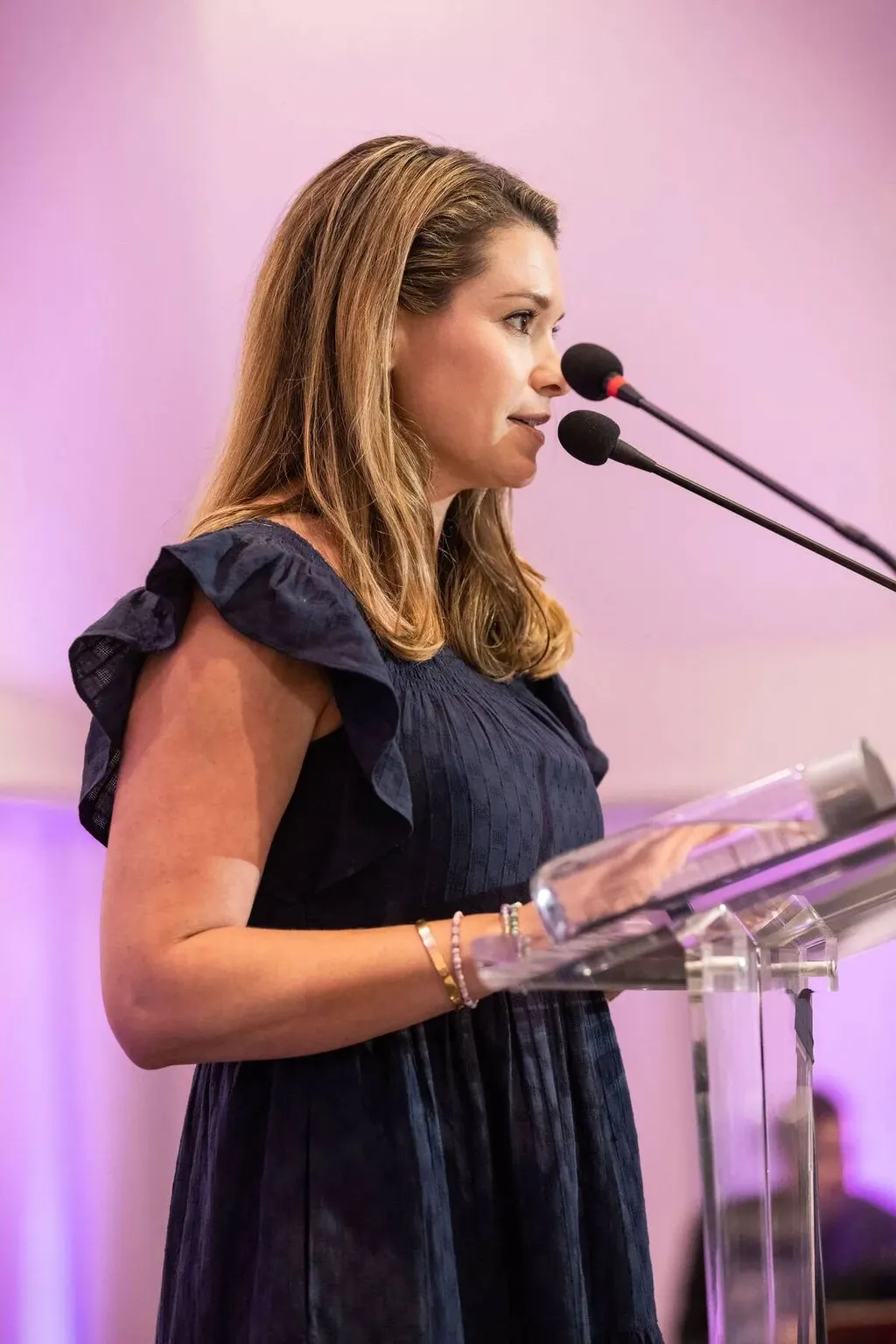 Woman speaking at a glass podium under pink event lighting at the Nadel University conference, photographed by Marquee Visuals.