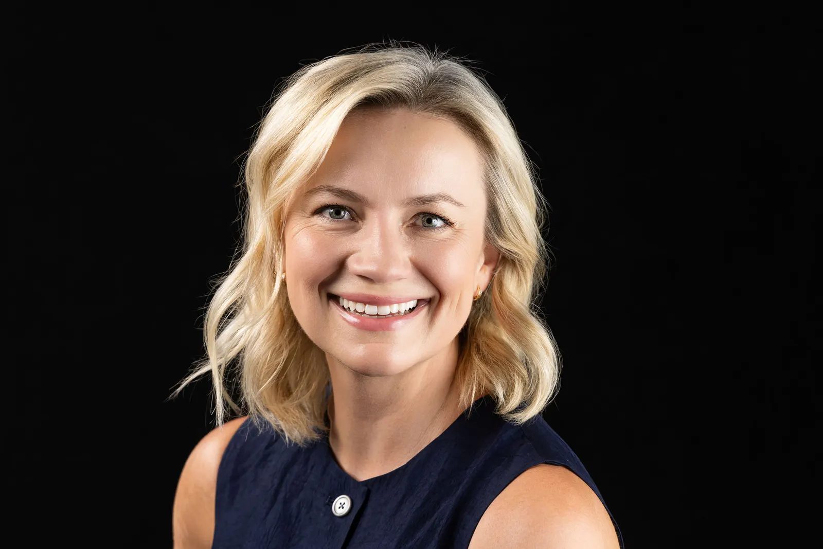 Executive headshot of a smiling blonde woman in a navy sleeveless top on a black studio backdrop, photographed by Marquee Visuals in Los Angeles.