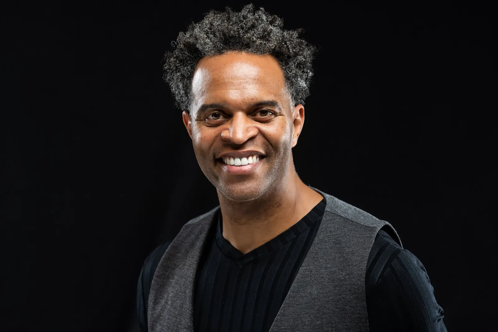 Corporate headshot of a smiling man with curly gray hair in a dark vest over a black turtleneck on a black studio backdrop, by Marquee Visuals in Los Angeles.
