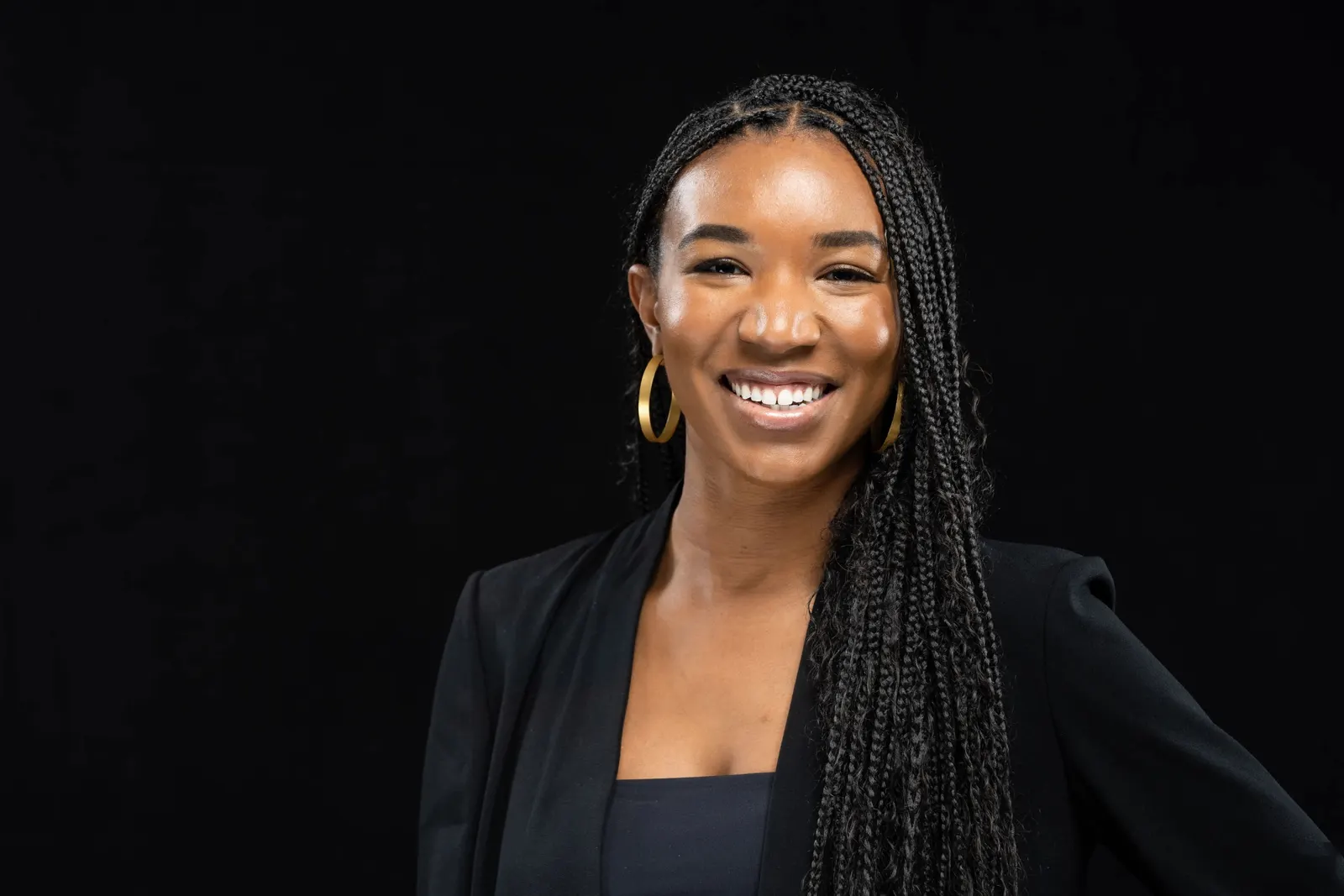 Corporate headshot of a smiling woman with braided hair in a black blazer and gold earrings on a black studio backdrop, by Marquee Visuals in Los Angeles.