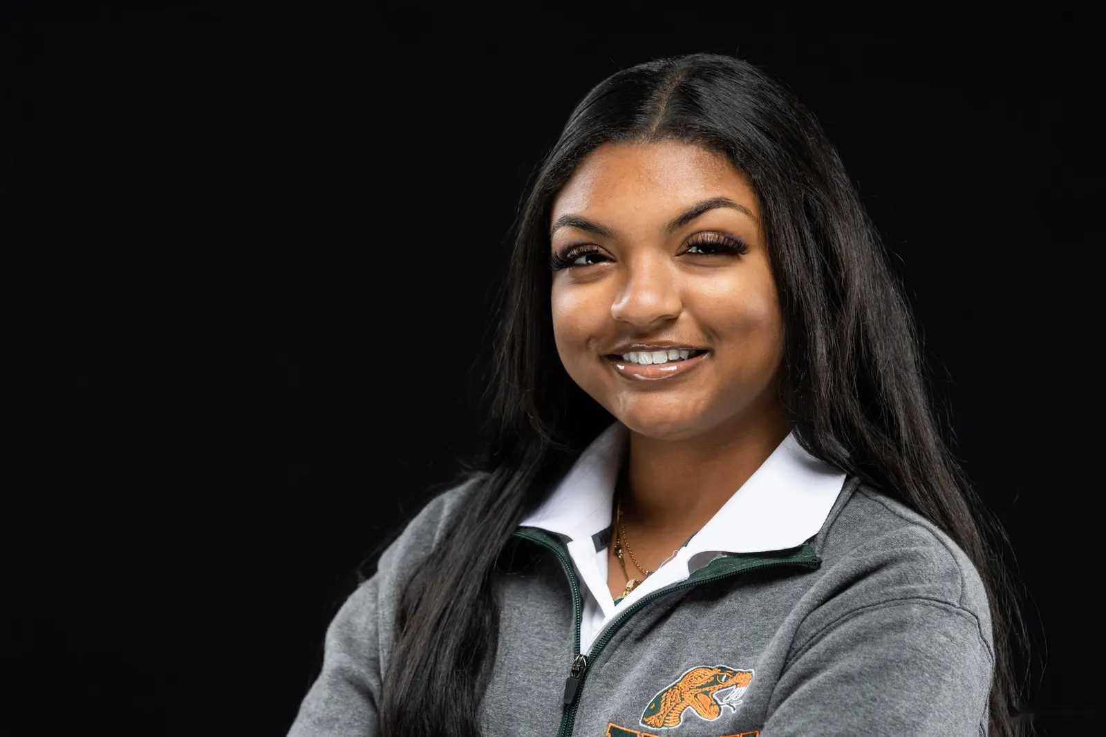 Corporate headshot of a smiling woman with wavy dark hair, a gold necklace, and a black top on a black studio backdrop, by Marquee Visuals in Los Angeles.