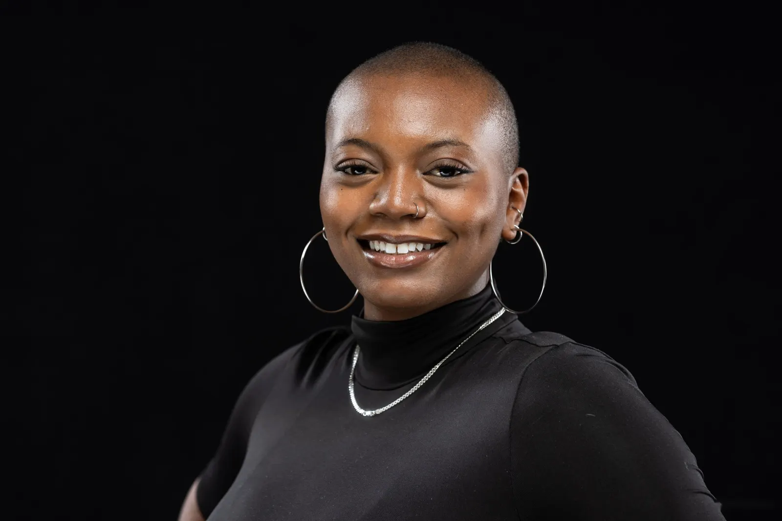 Corporate headshot of a smiling woman with short buzzed hair and hoop earrings in a black top on a black studio backdrop, by Marquee Visuals in Los Angeles.
