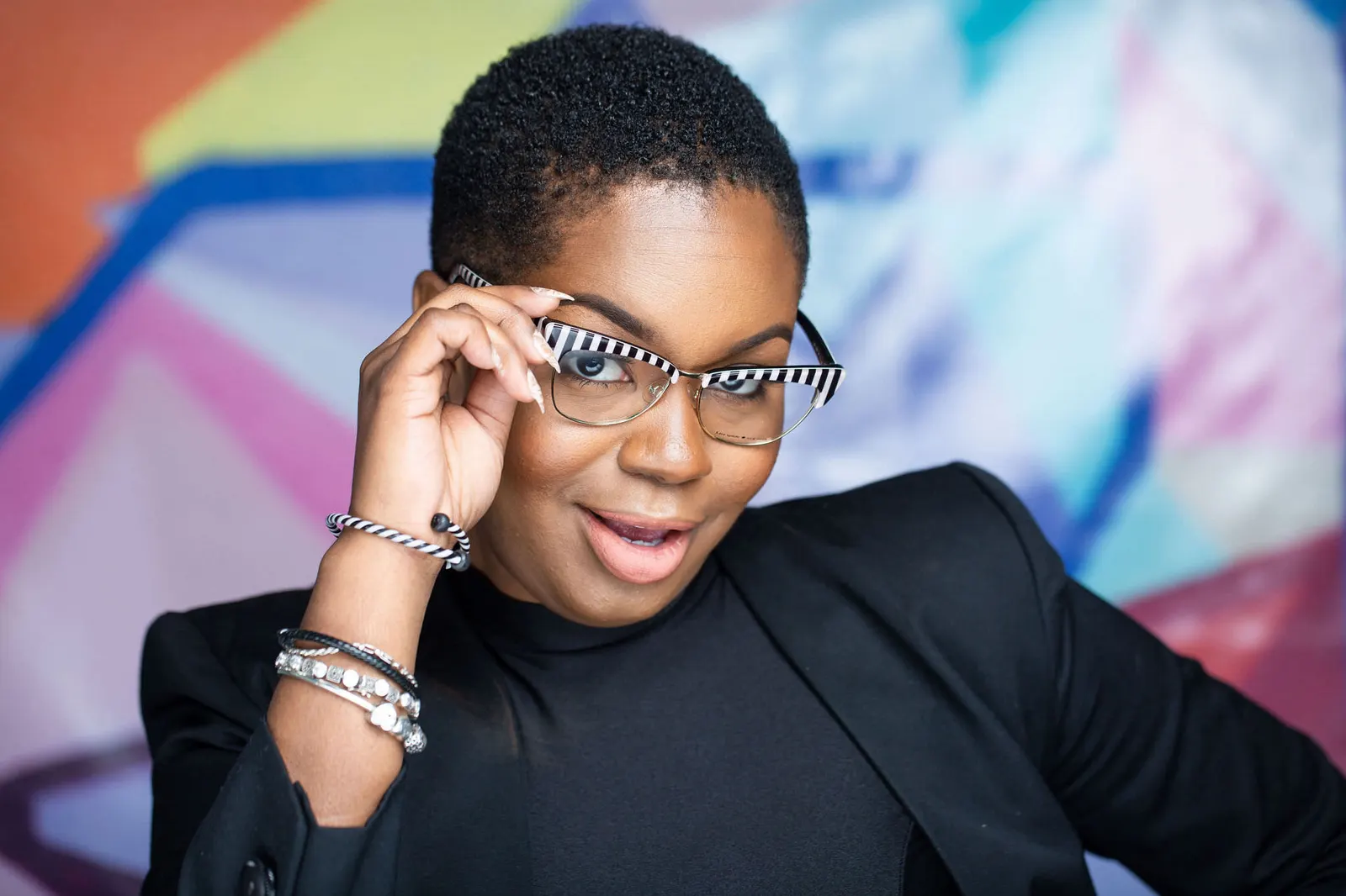 Candid headshot of a smiling woman adjusting her glasses in a black top against a colorful mural backdrop, by Marquee Visuals in Los Angeles.