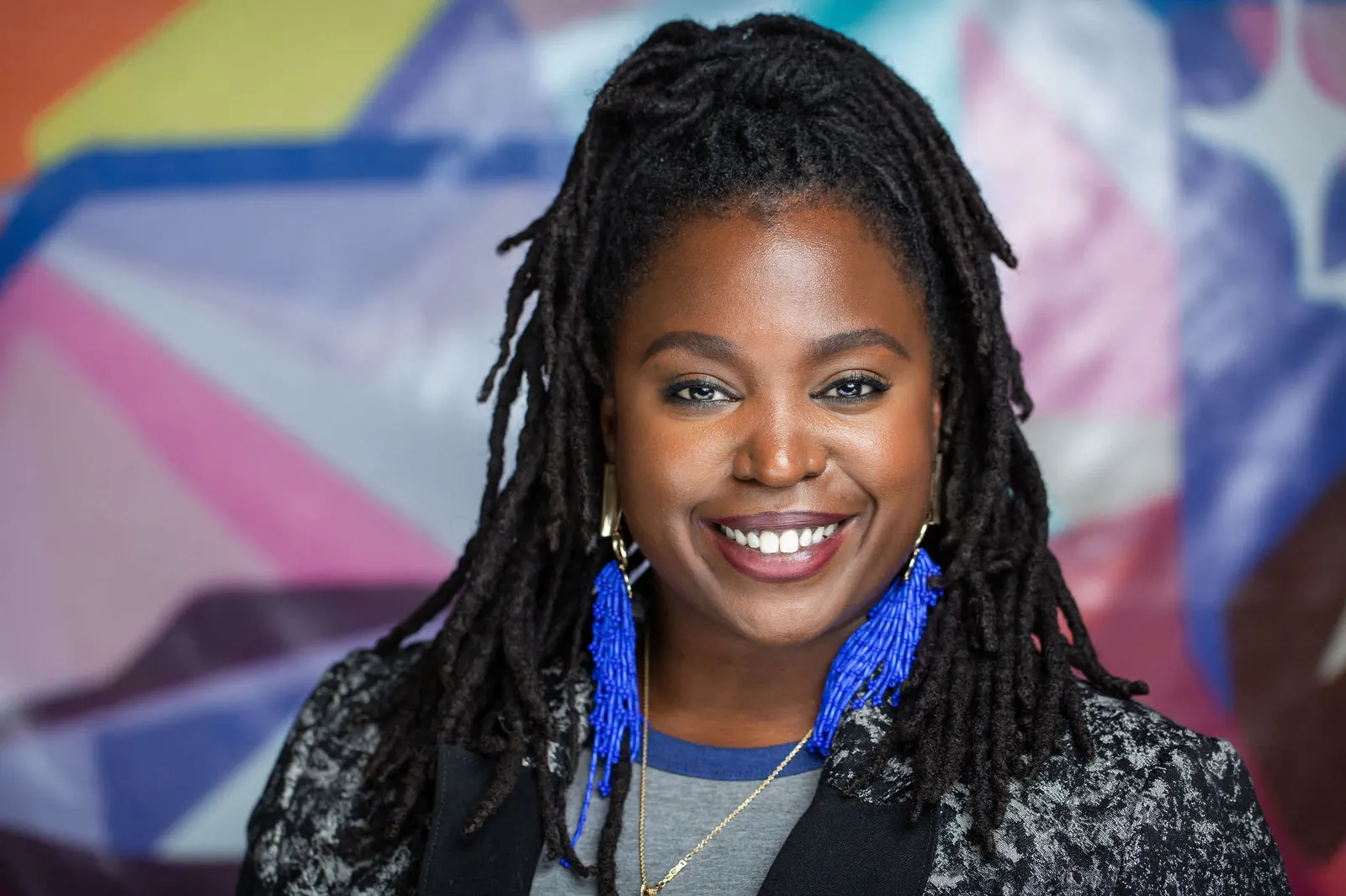 Corporate headshot of a smiling woman with braided hair and blue beaded earrings against a colorful mural backdrop, by Marquee Visuals in Los Angeles.