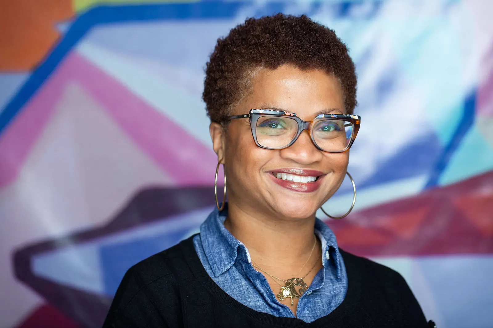 Corporate headshot of a smiling woman with curly hair and glasses in a denim shirt against a colorful mural backdrop, by Marquee Visuals in Los Angeles.