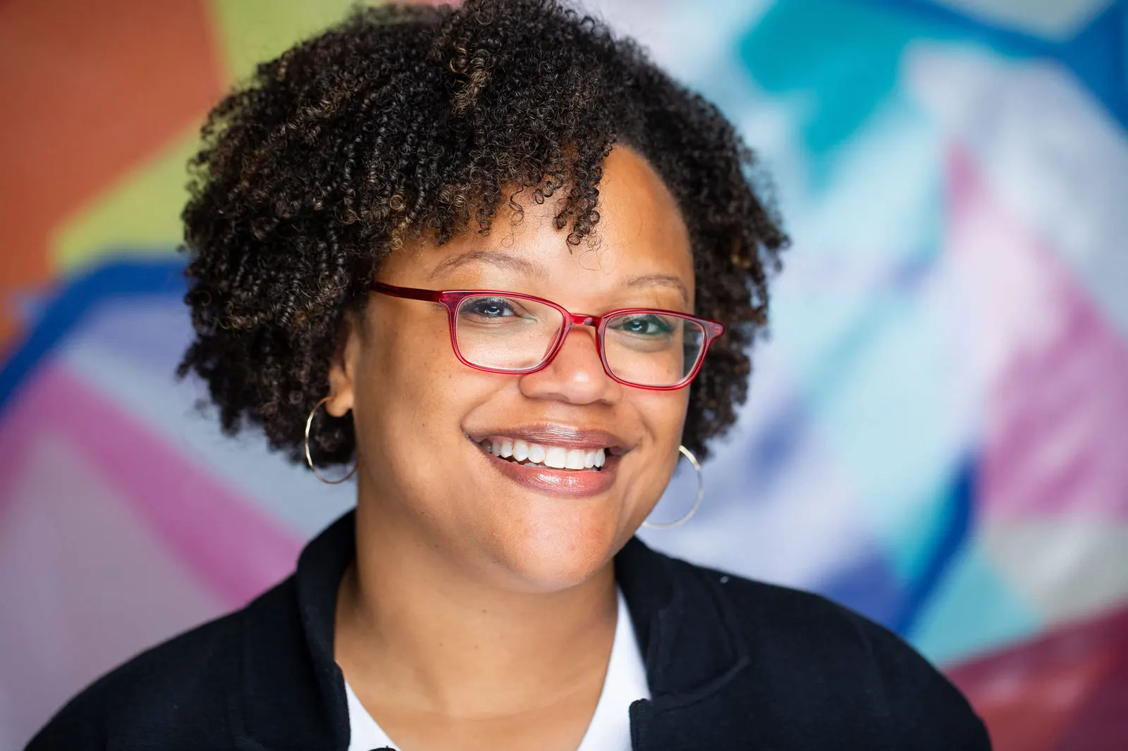 Corporate headshot of a smiling woman with red glasses and a black cardigan against a colorful mural backdrop, photographed by Marquee Visuals in Los Angeles.
