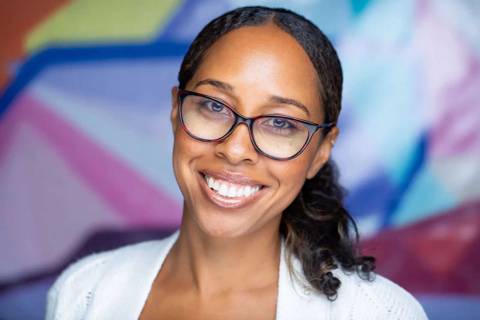 Corporate headshot of a smiling woman with glasses and curly hair in a white blazer against a colorful mural backdrop, by Marquee Visuals in Los Angeles.