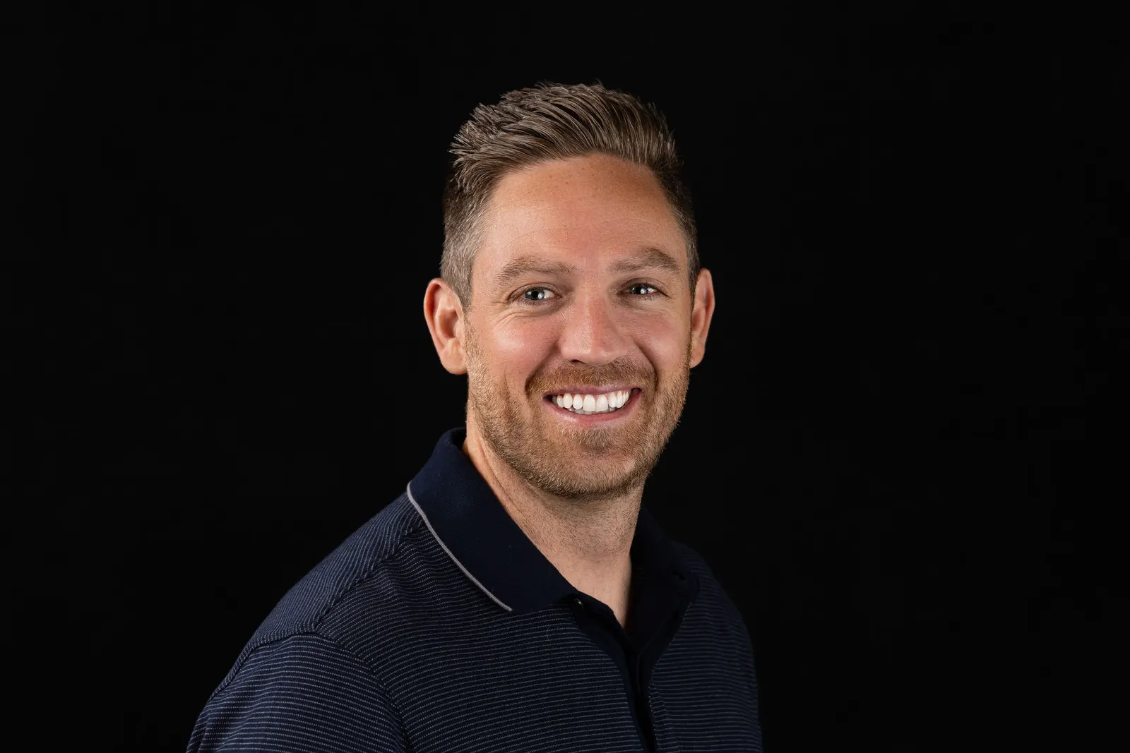 Corporate headshot of a smiling man with short blonde hair in a navy polo shirt on a black studio backdrop, photographed by Marquee Visuals in Los Angeles.