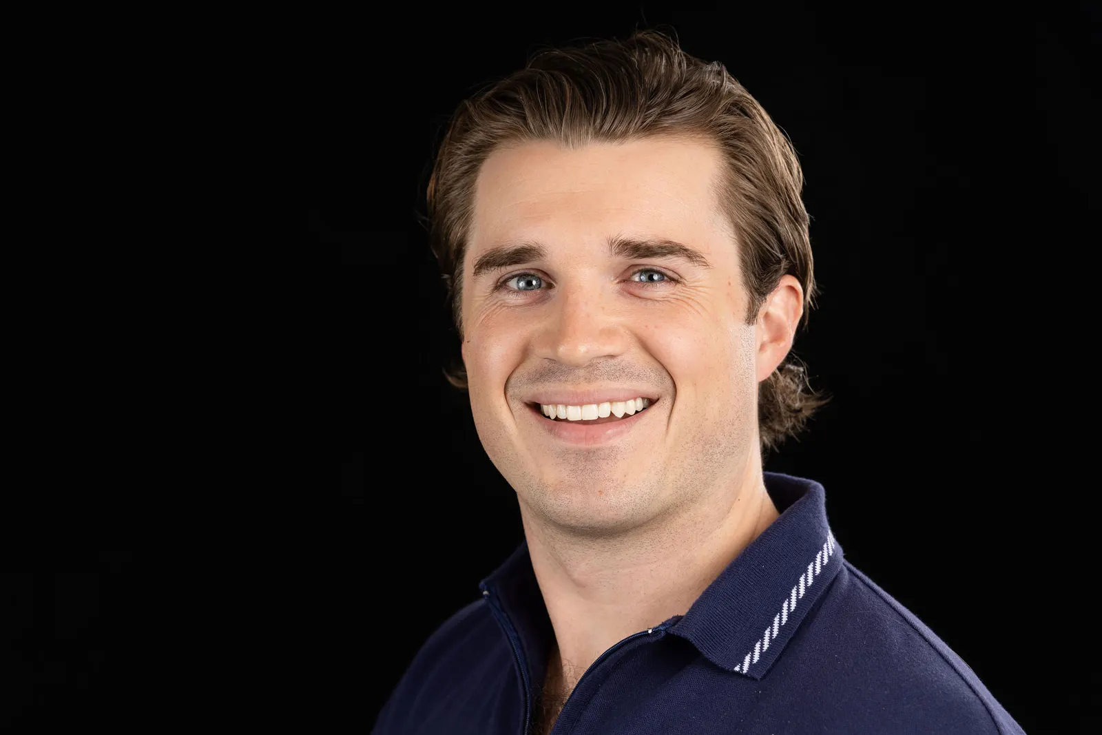 Corporate headshot of a smiling man with short brown hair in a navy striped polo on a black studio backdrop, photographed by Marquee Visuals in Los Angeles.
