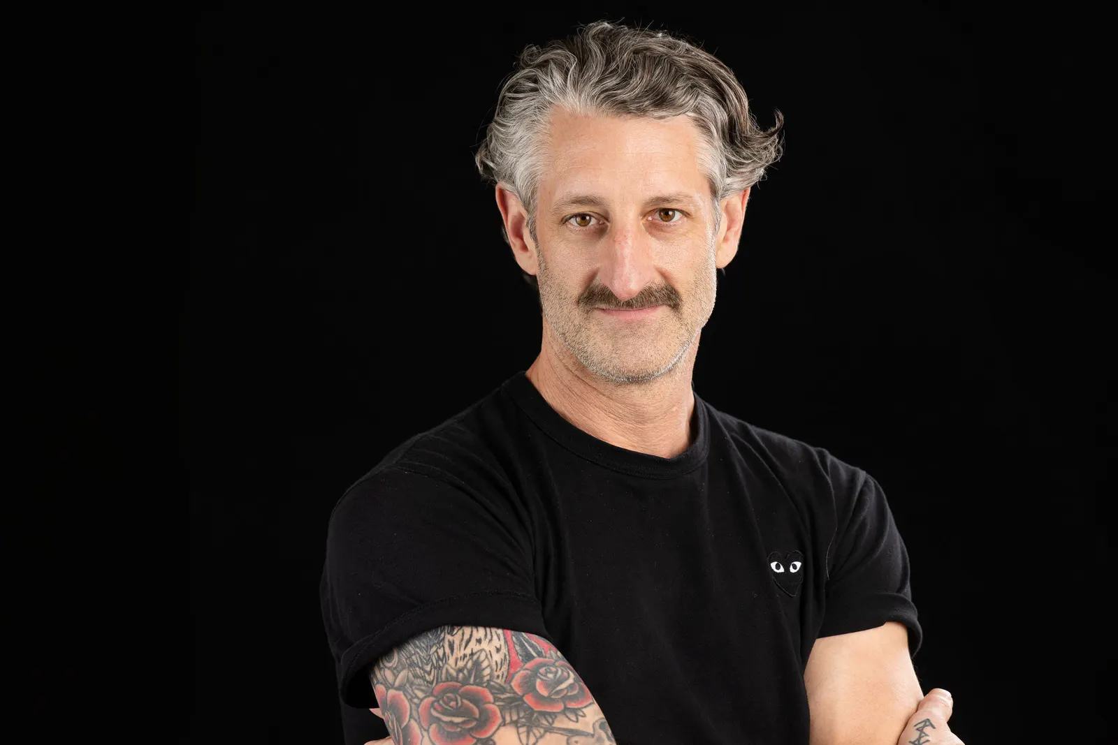 Corporate headshot of a gray-haired man with a mustache, tattoos, and a black t-shirt, arms crossed, on a black studio backdrop, by Marquee Visuals in Los Angeles.