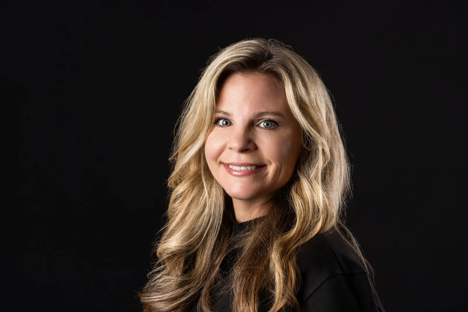 Corporate headshot of a smiling long-haired blonde woman on a black studio backdrop, photographed by Marquee Visuals in Los Angeles.