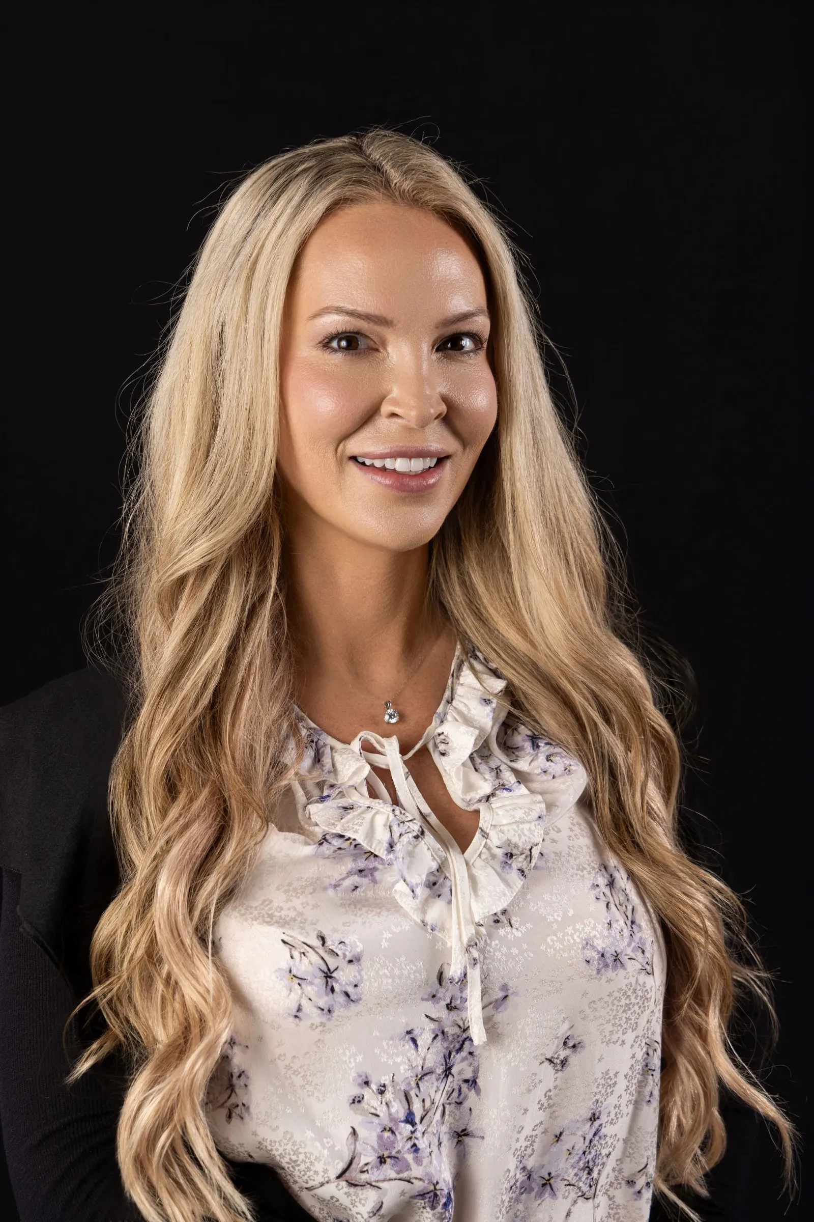 Corporate headshot of a smiling blonde woman in a floral blouse on a black studio backdrop, photographed by Marquee Visuals in Los Angeles.