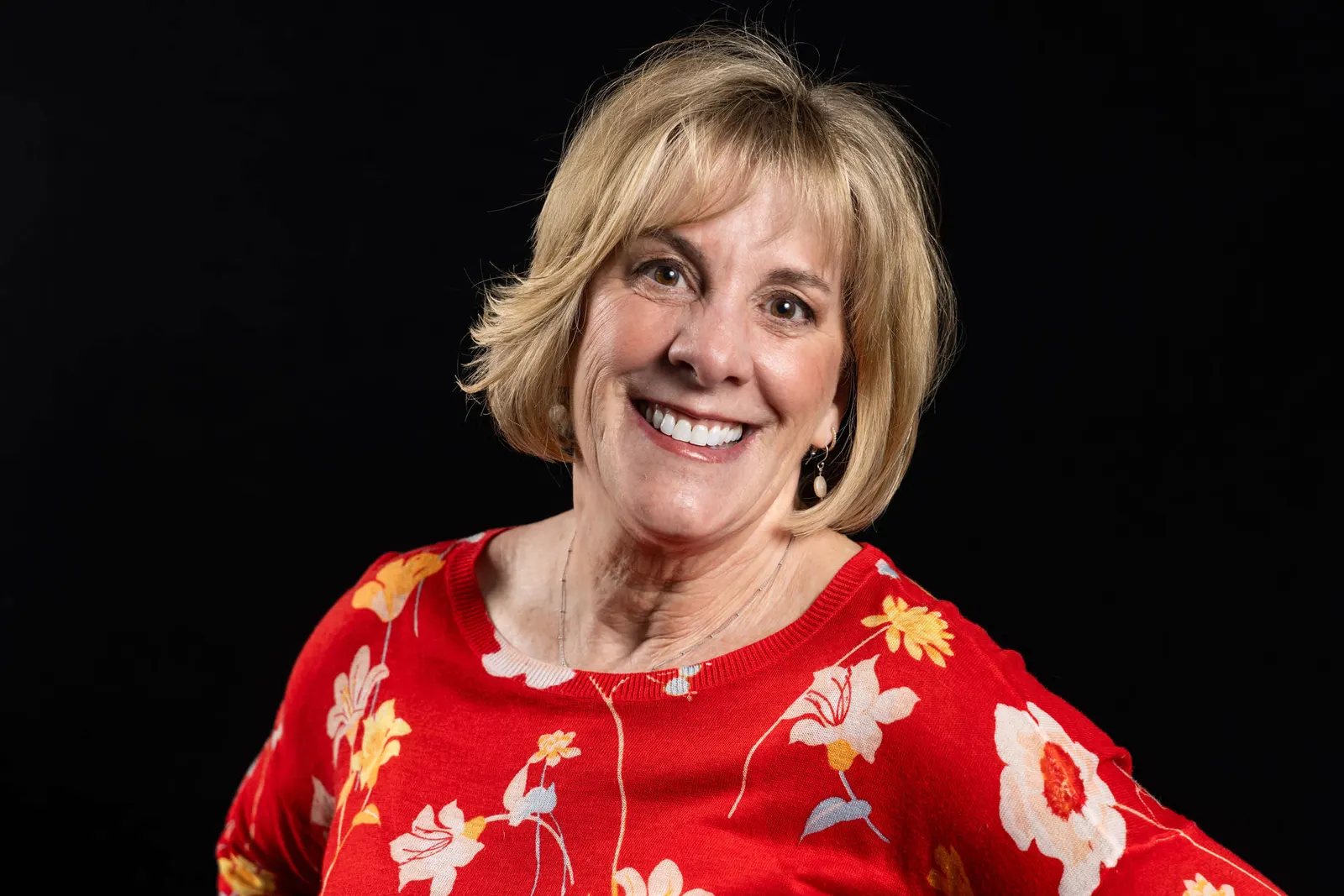 Corporate headshot of a smiling curly-haired woman in a bright red floral top on a black studio backdrop, photographed by Marquee Visuals in Los Angeles.
