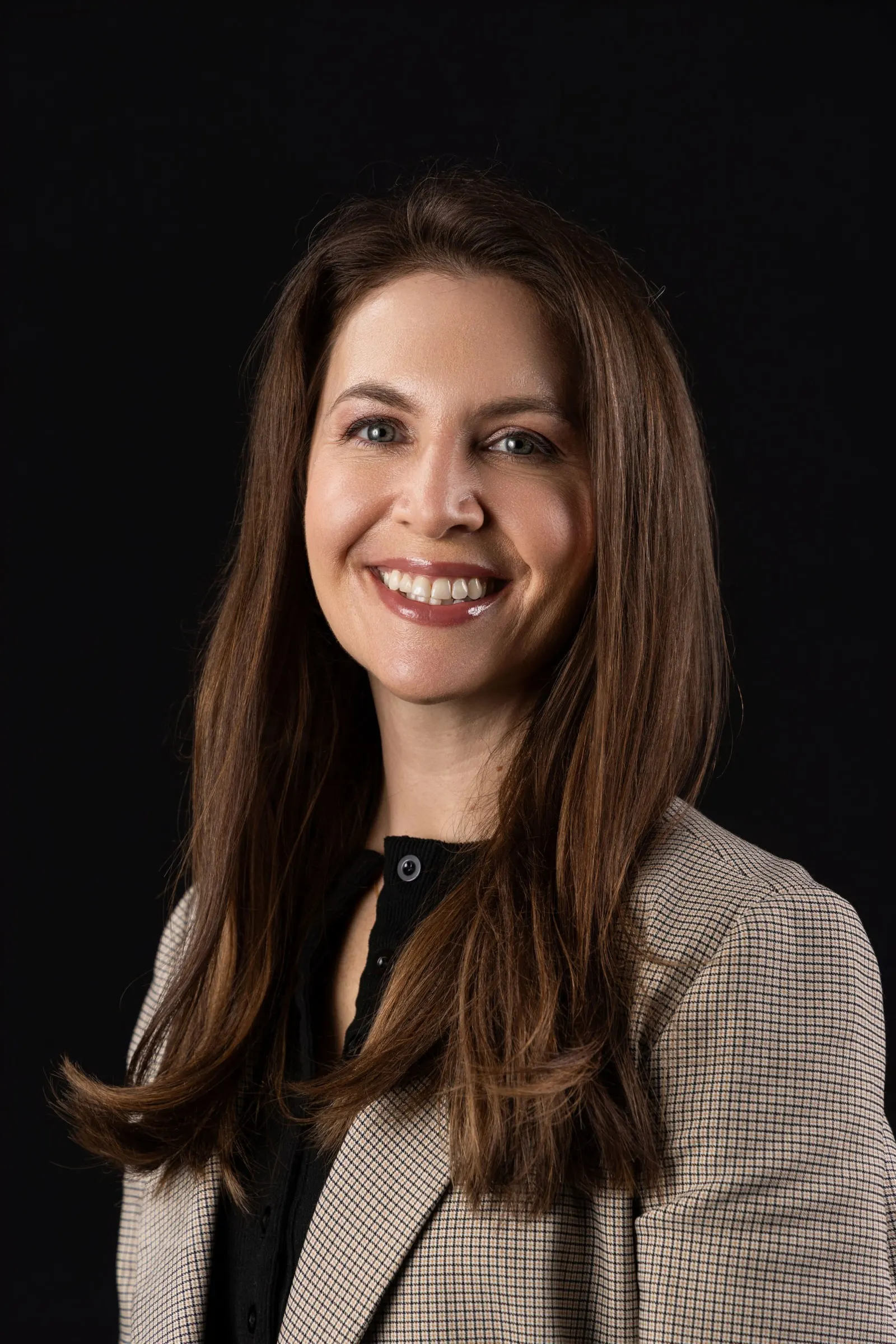 Corporate headshot of a smiling brunette woman in a tan blazer on a black studio backdrop, photographed by Marquee Visuals in Los Angeles.