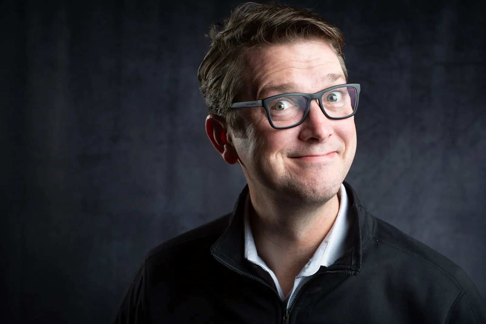 Corporate headshot of a man with glasses and a wry smile in a dark shirt on a dark gray studio backdrop, photographed by Marquee Visuals in Los Angeles.