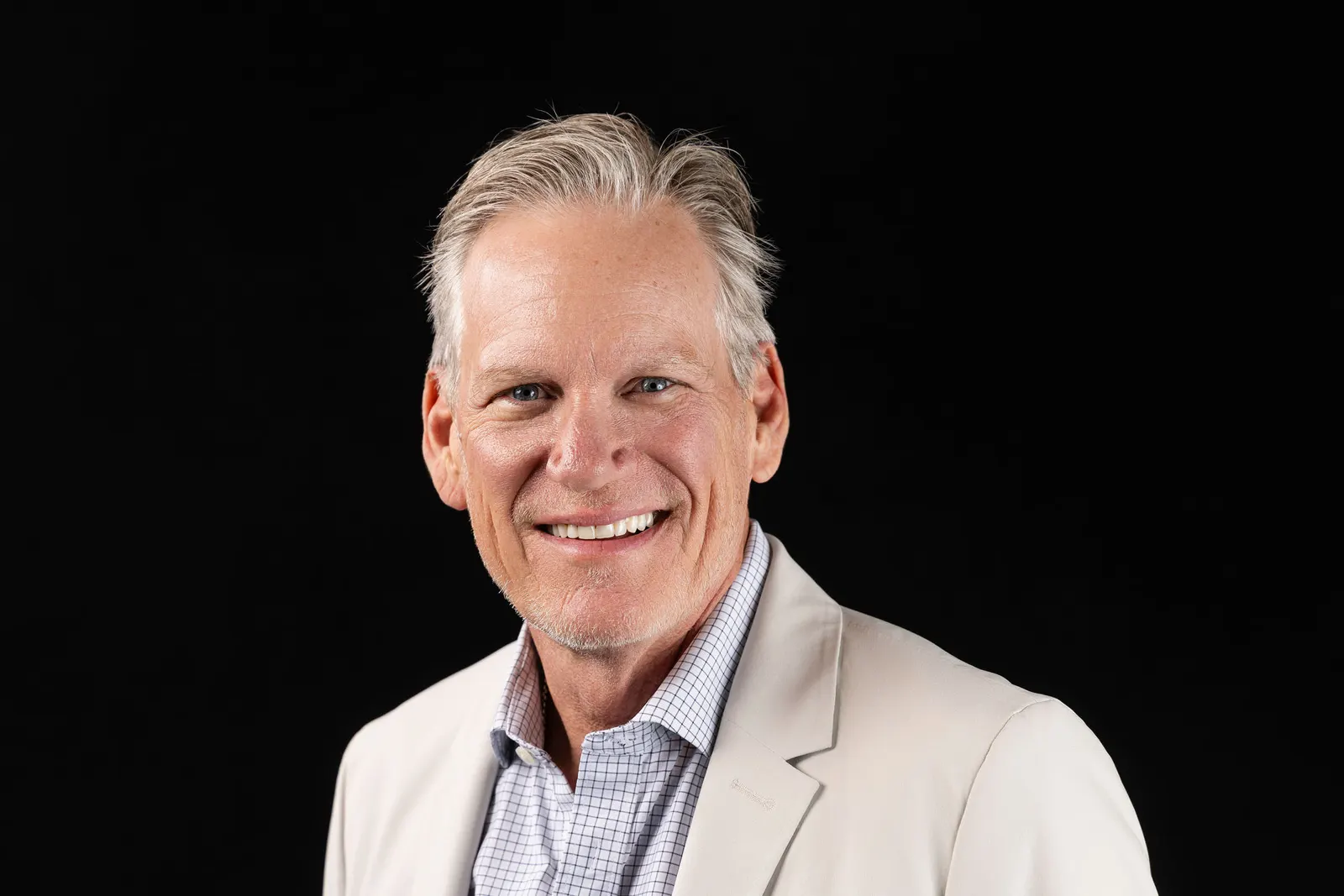 Corporate headshot of a smiling gray-haired executive in a tan blazer and checked shirt on a black backdrop, photographed by Marquee Visuals in Los Angeles.