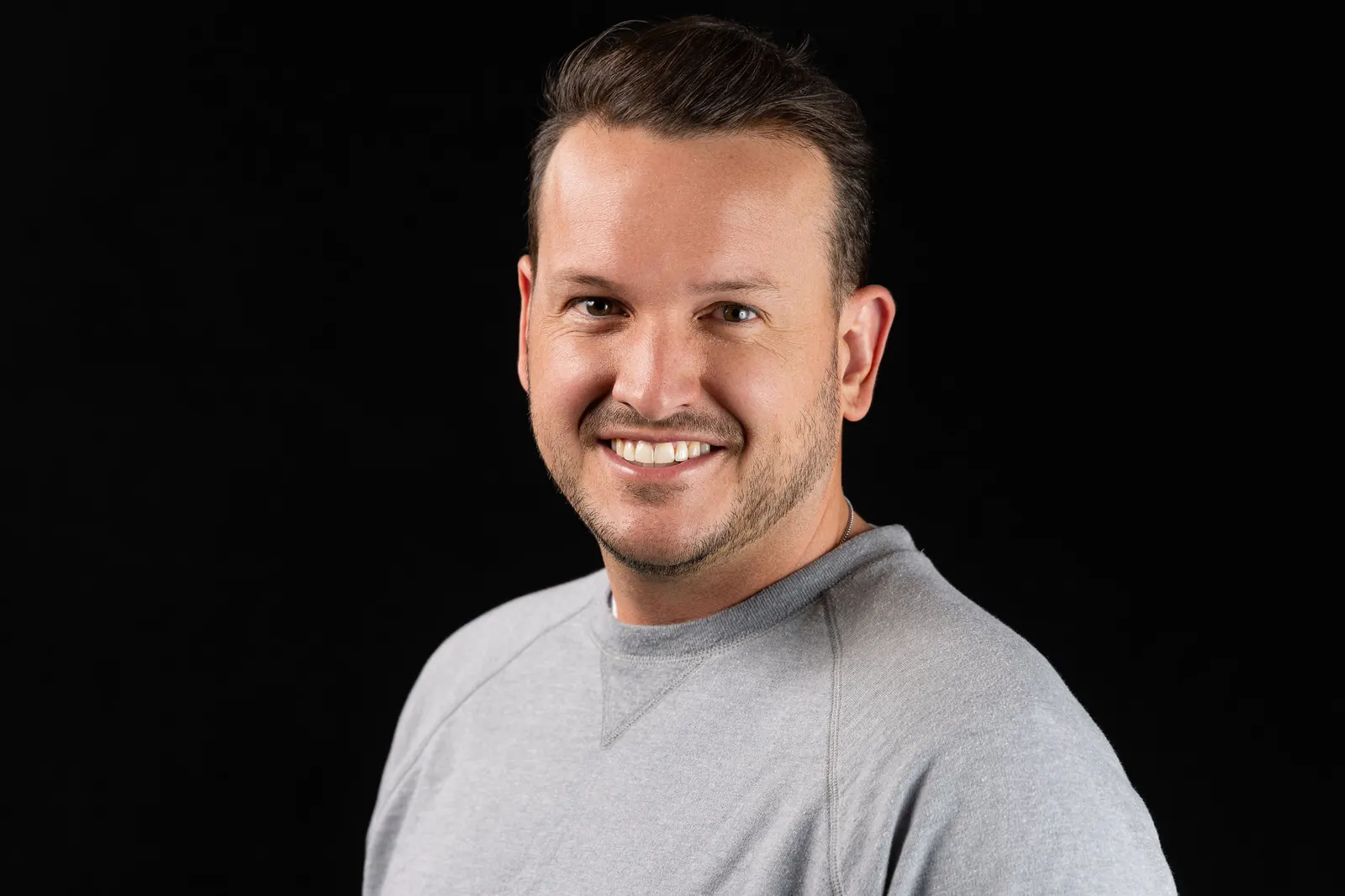 Corporate headshot of a smiling man in a light gray crewneck on a black backdrop, photographed by Marquee Visuals in Los Angeles.