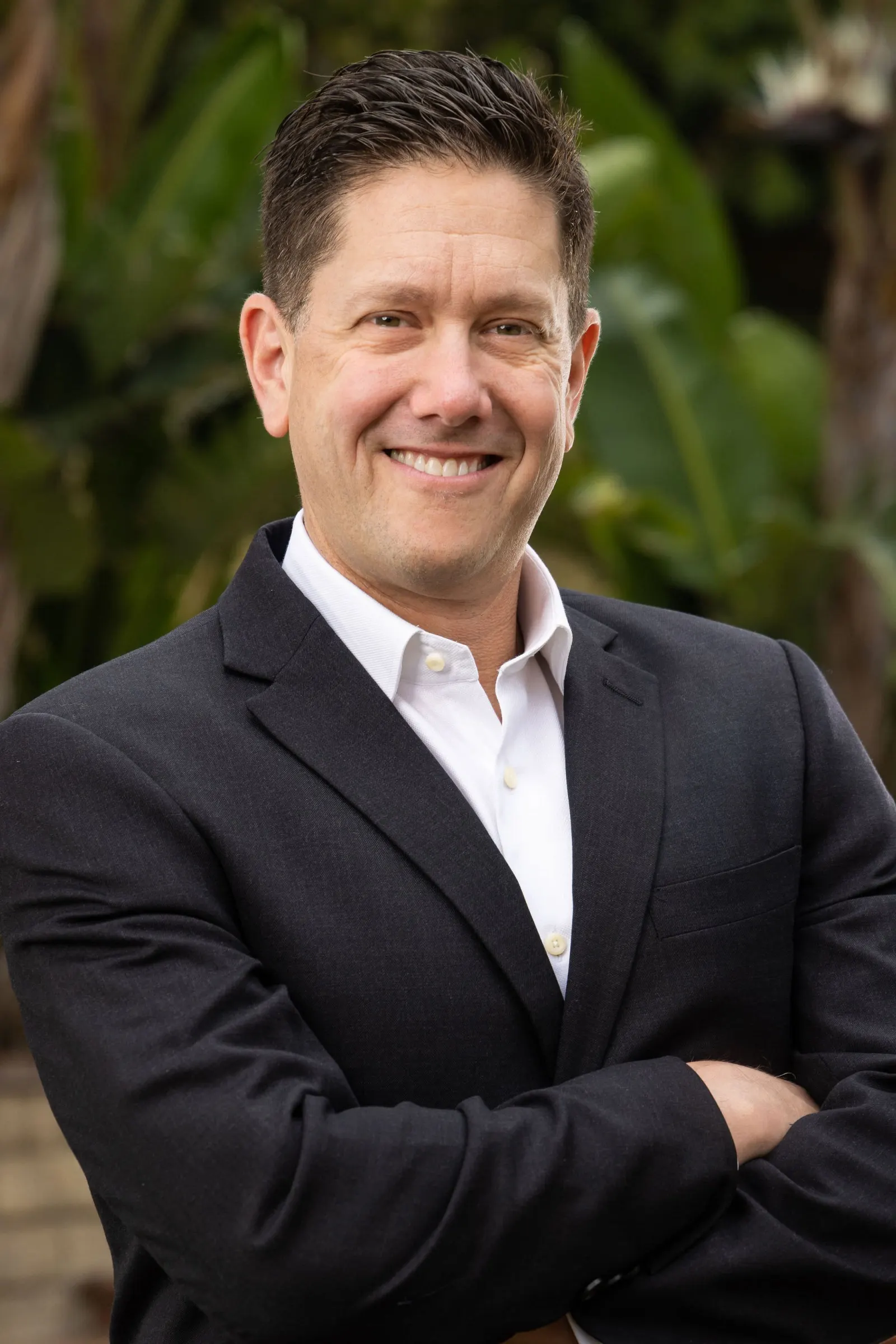 Executive headshot of a man with glasses in a dark suit, arms crossed, photographed outdoors with a soft green bokeh backdrop, by Marquee Visuals in Los Angeles.