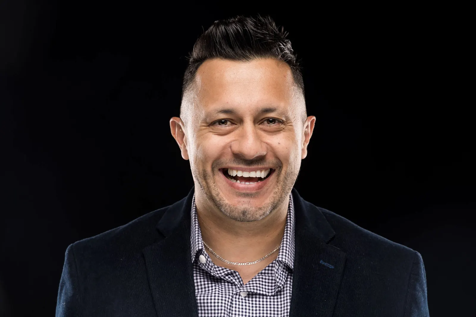 Corporate headshot of a smiling man with dark hair in a navy checked shirt on a black studio backdrop, photographed by Marquee Visuals in Los Angeles.