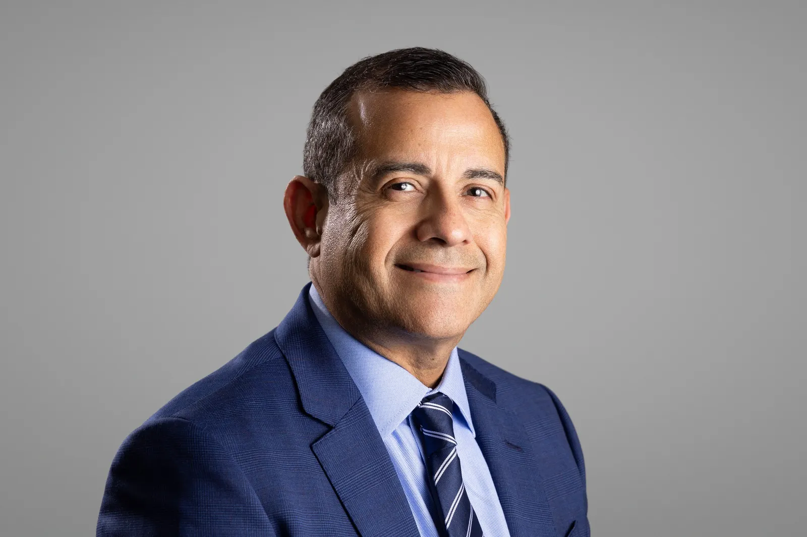 Corporate headshot of a smiling man in a navy suit and tie on a light gray studio backdrop, photographed by Marquee Visuals in Los Angeles.