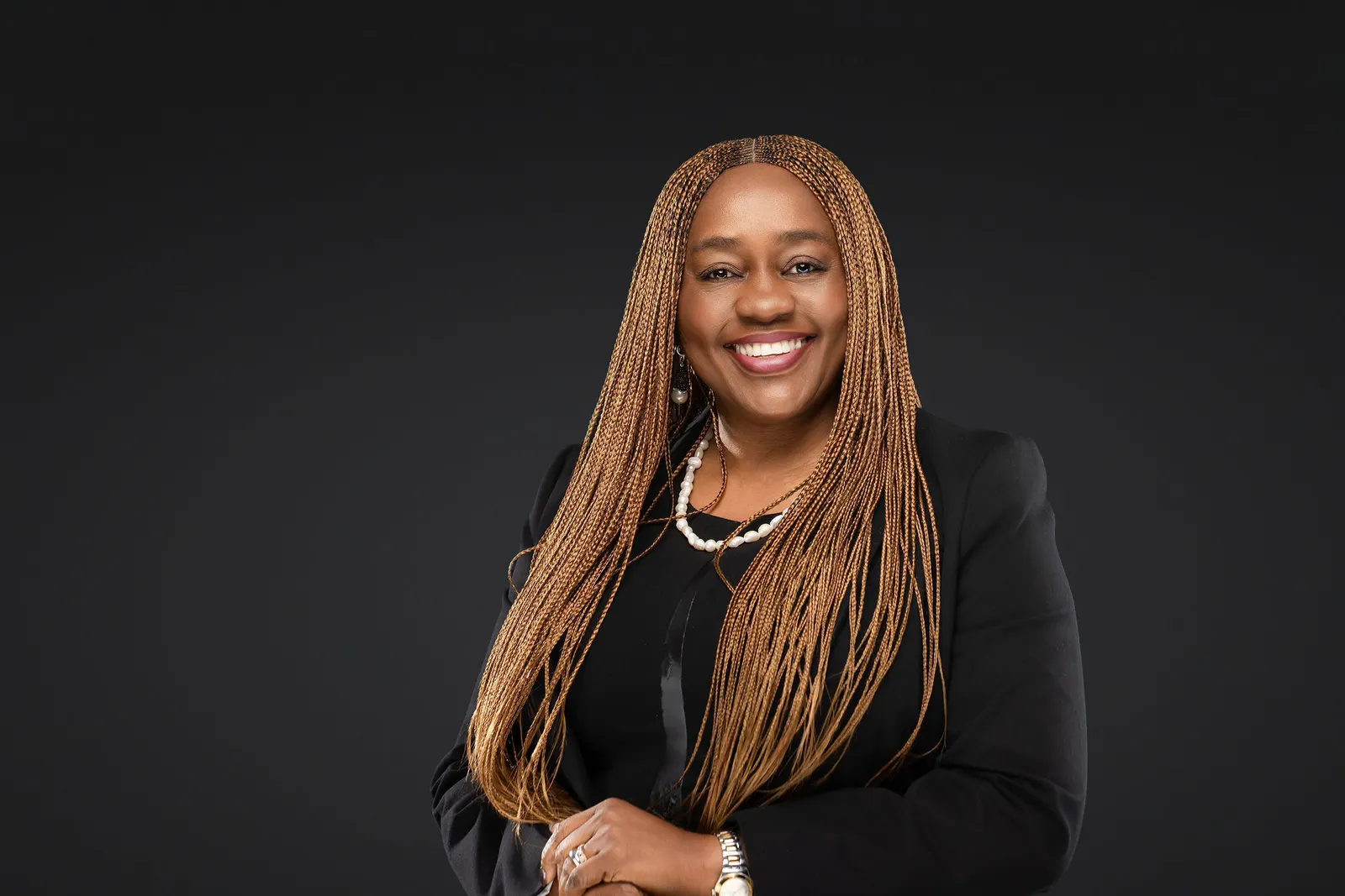 Executive headshot of a woman with long braided hair and a black blazer, arms crossed, on a soft gradient gray backdrop, by Marquee Visuals in Los Angeles.