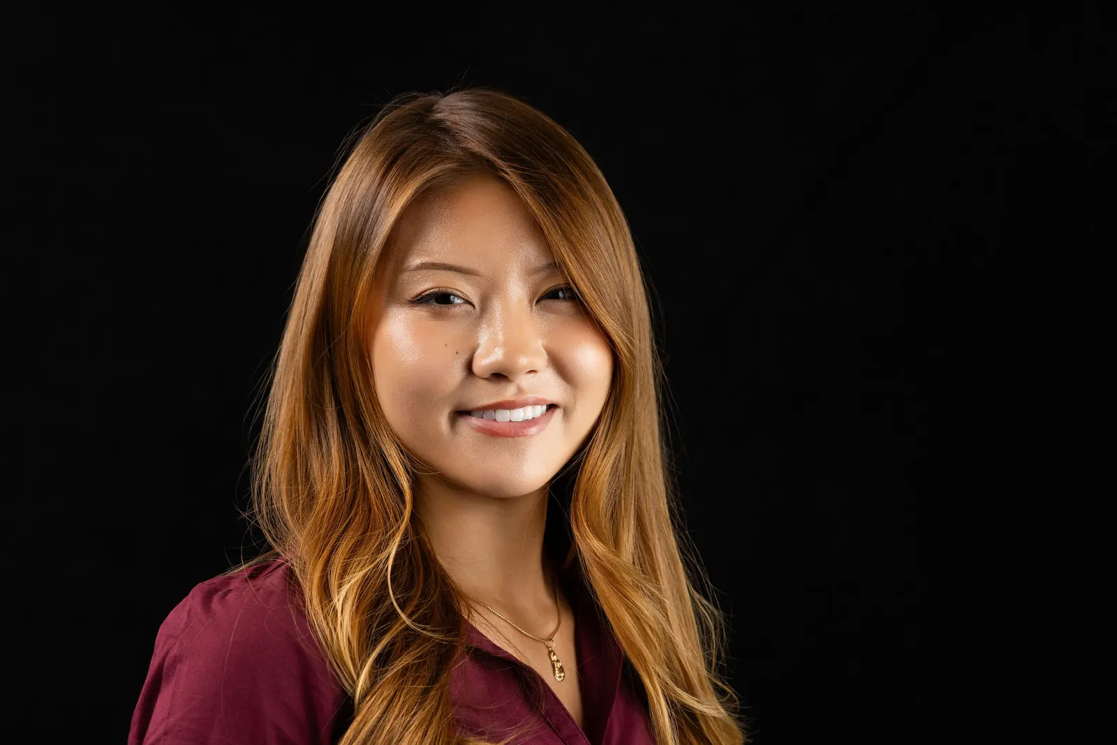 Corporate headshot of a smiling woman with auburn hair in a maroon blouse on a black studio backdrop, photographed by Marquee Visuals in Los Angeles.