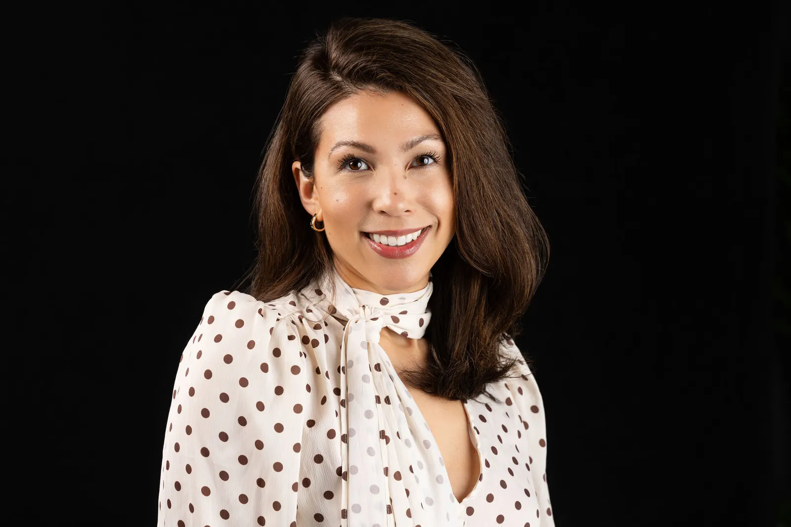 Corporate headshot of a smiling brunette woman in a white polka-dot blouse with a bow collar on a black backdrop, by Marquee Visuals in Los Angeles.