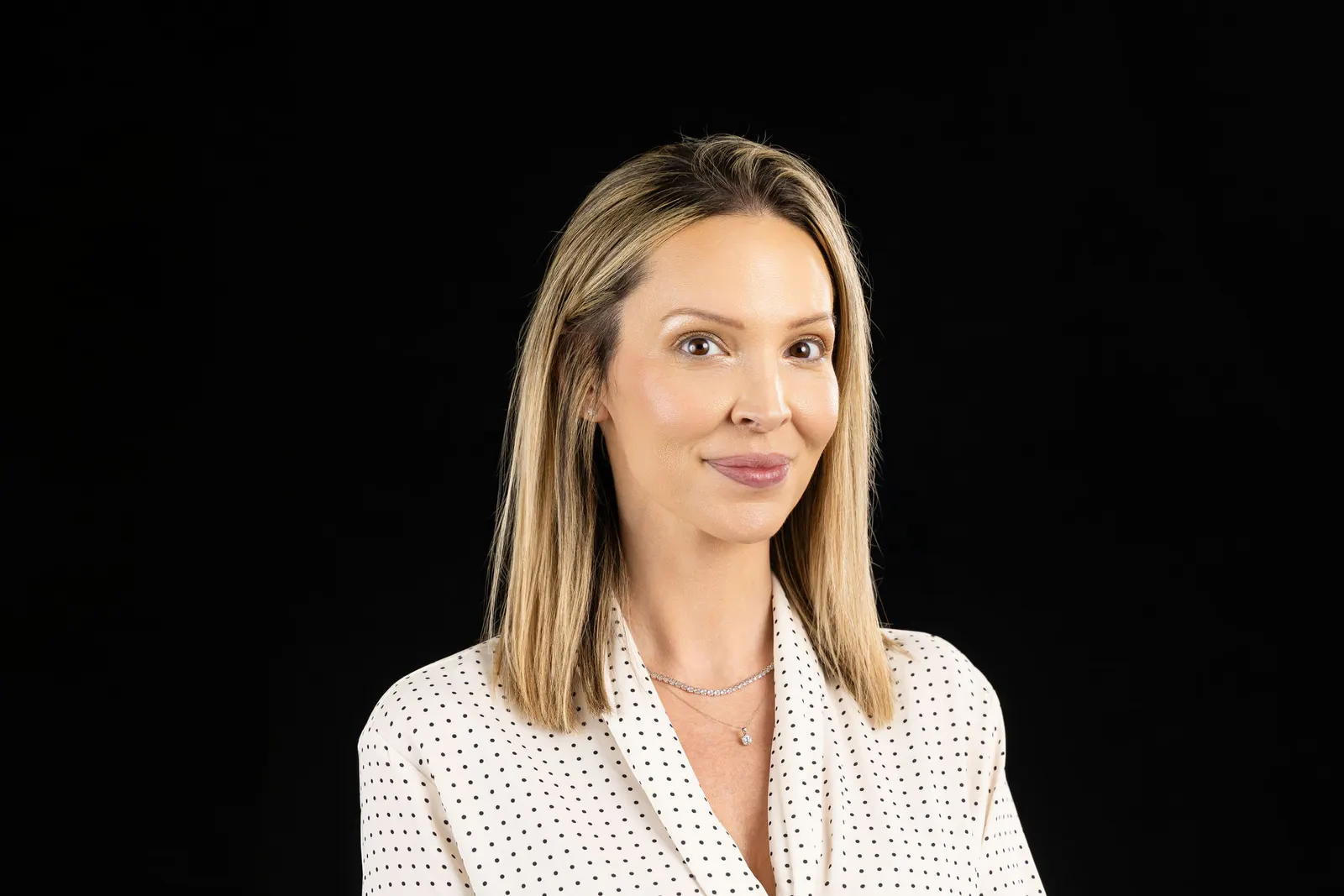Corporate headshot of a smiling blonde woman in a white polka-dot blouse on a black studio backdrop, photographed by Marquee Visuals in Los Angeles.