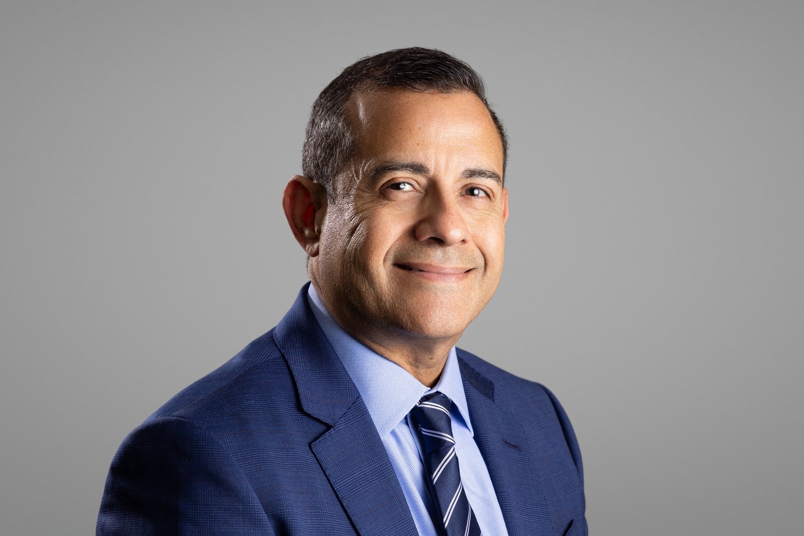 Corporate headshot of a smiling man in a navy suit and tie on a light gray studio backdrop, photographed by Marquee Visuals in Los Angeles.